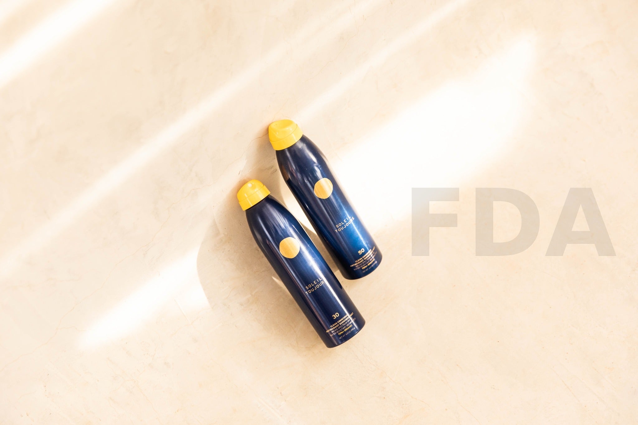 Two Soleil Toujours Clean Conscious Sunscreen Mists with subtle FDA watermark on stone wall — elegant, science-led SPF visual